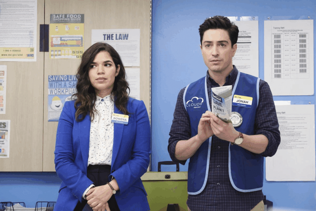 Uma foto de cena de Superstore com dois funcionários da Cloud 9, Rowena e Jonah, em pé na sala de descanso ou escritório da loja. Rowena, à esquerda, está de blazer azul sobre uma camisa estampada e calças pretas, olhando para o lado. Jonah, à direita, está de colete Cloud 9 azul sobre uma camisa xadrez, segurando e olhando para um folheto. O fundo é uma parede azul com vários pôsteres e quadros de avisos.