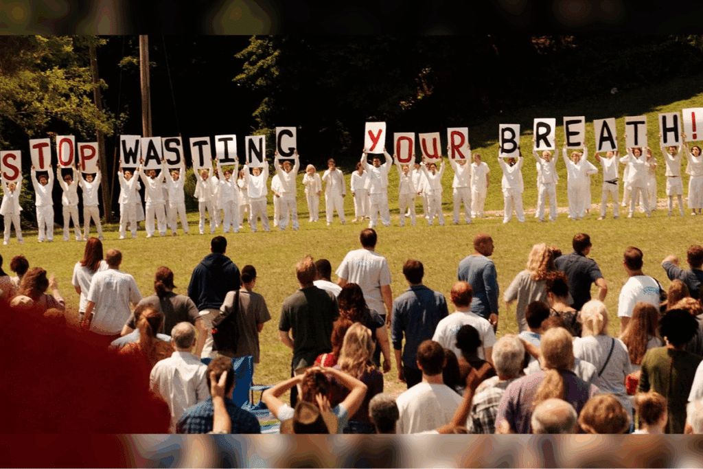 Uma multidão de costas observa um grupo de pessoas vestidas inteiramente de branco, alinhadas em um gramado. As pessoas de branco seguram placas que formam a frase "STOP WASTING YOUR BREATH!" (Pare de desperdiçar seu fôlego!)