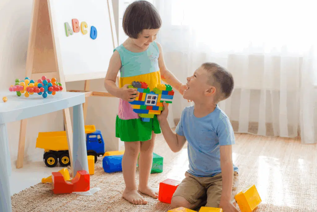 Menina com cabelo curto, usando vestido listrado azul, amarelo e verde dando brinquedo de blocos para um menino com cabelo curto, usando camiseta azul e bermuda bege em quarto com brinquedos 