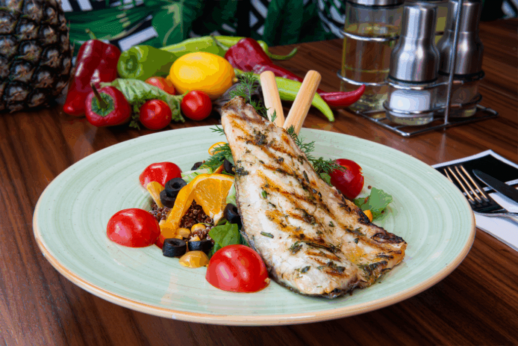 Filé de peixe grelhado com tomate-cereja, pedaços de laranja, folhas verdes, azeitona preta e alecrim