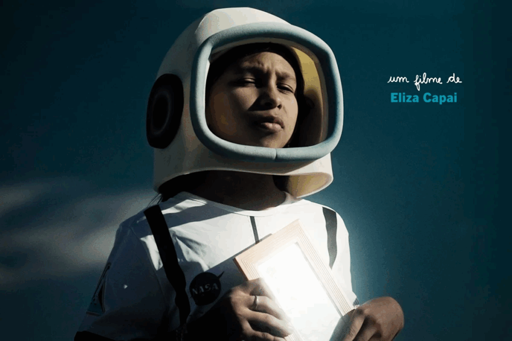 Menina usando capacete de astronauta e uniforme de astronauta branco com detalhes em preto, segurando um porta retrato junto ao corpo em fundo azul 