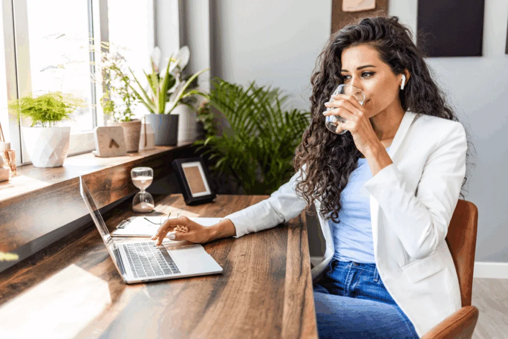 Mulher com cabelo cacheado longo, usando camiseta e blazer branco e calça jeans sentada em mesa de escritório com notebook na mesa bebendo água em copo de vidro