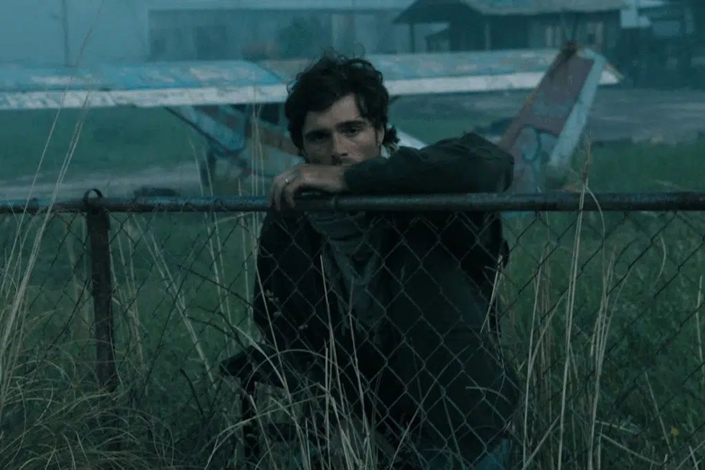 Jacob Elordi agachado em uma cerca com grama em cena de "Ponto sem Retorno"