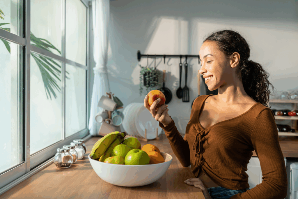 Mulher sorridente segura uma maçã na cozinha, próxima a uma janela iluminada pelo sol e a uma fruteira repleta de frutas.