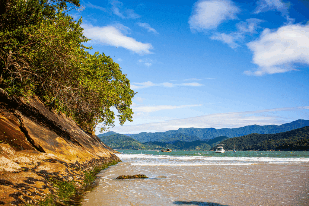 Uma praia de águas rasas e tranquilas, cercada por montanhas cobertas de vegetação. À esquerda, uma formação rochosa com árvores se estende até o mar. Pequenas embarcações aparecem ao longe, enquanto o céu azul, com algumas nuvens brancas, completa a paisagem.