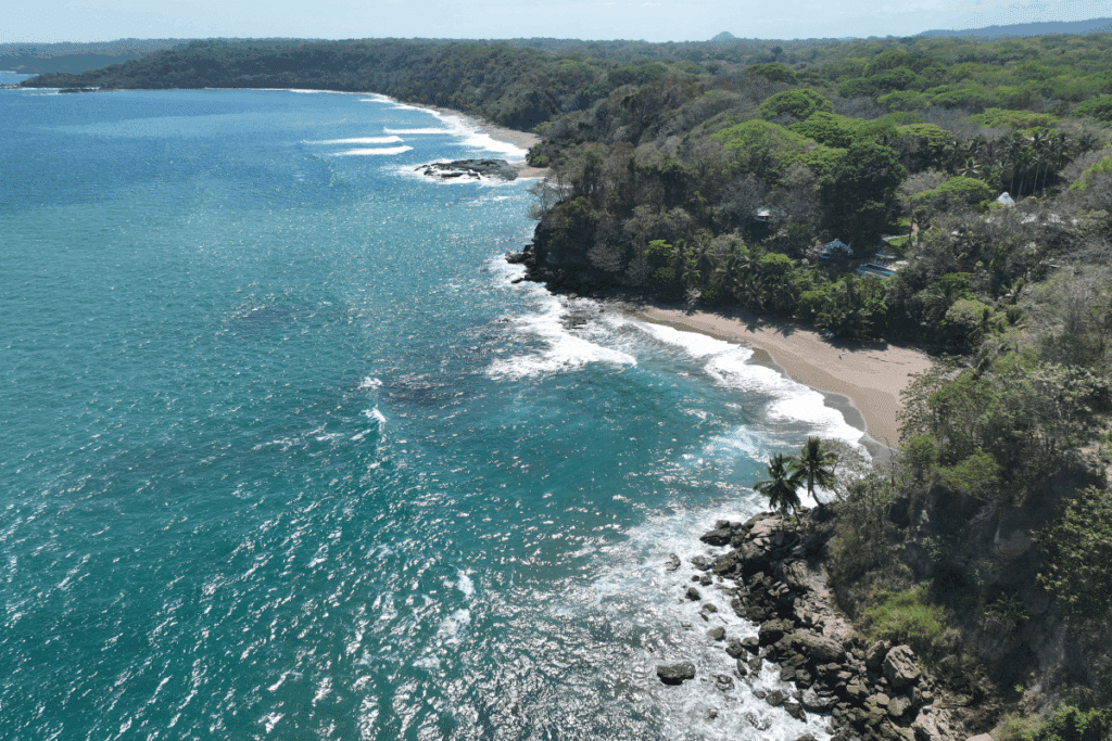 Vista aérea de uma enseada costeira cercada por floresta tropical densa e formações rochosas. Em primeiro plano, mar azul-esverdeado com reflexos solares e ondas suaves atingindo pequenas praias de areia escura