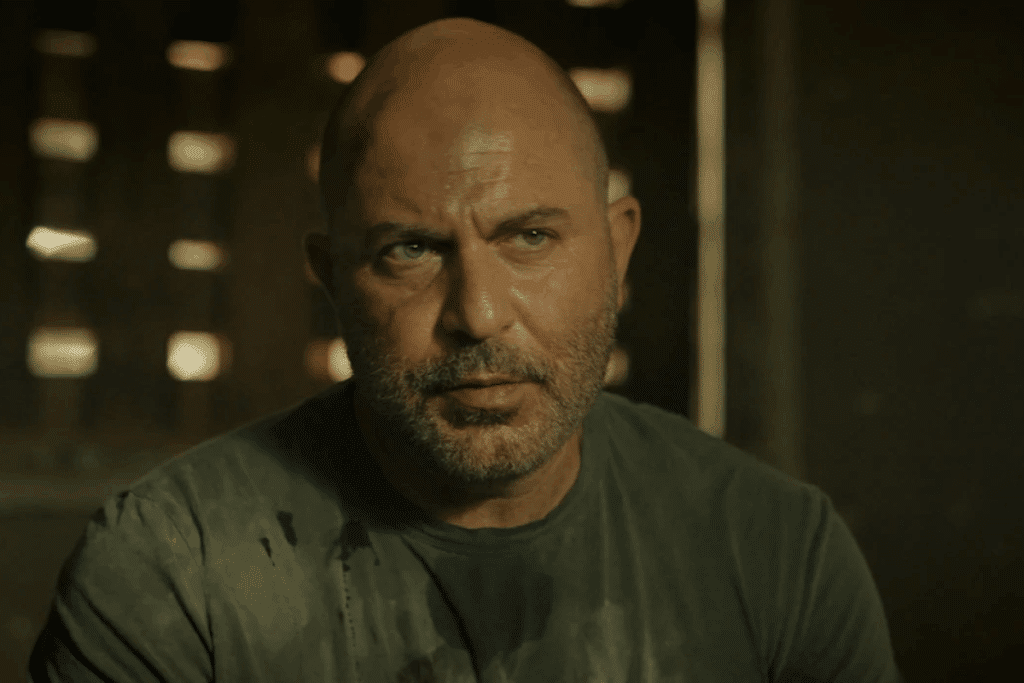 Personagem homem de Fauda com o rosto ferido e a camiseta encharcada, olhar tenso, dentro de um galpão escuro
