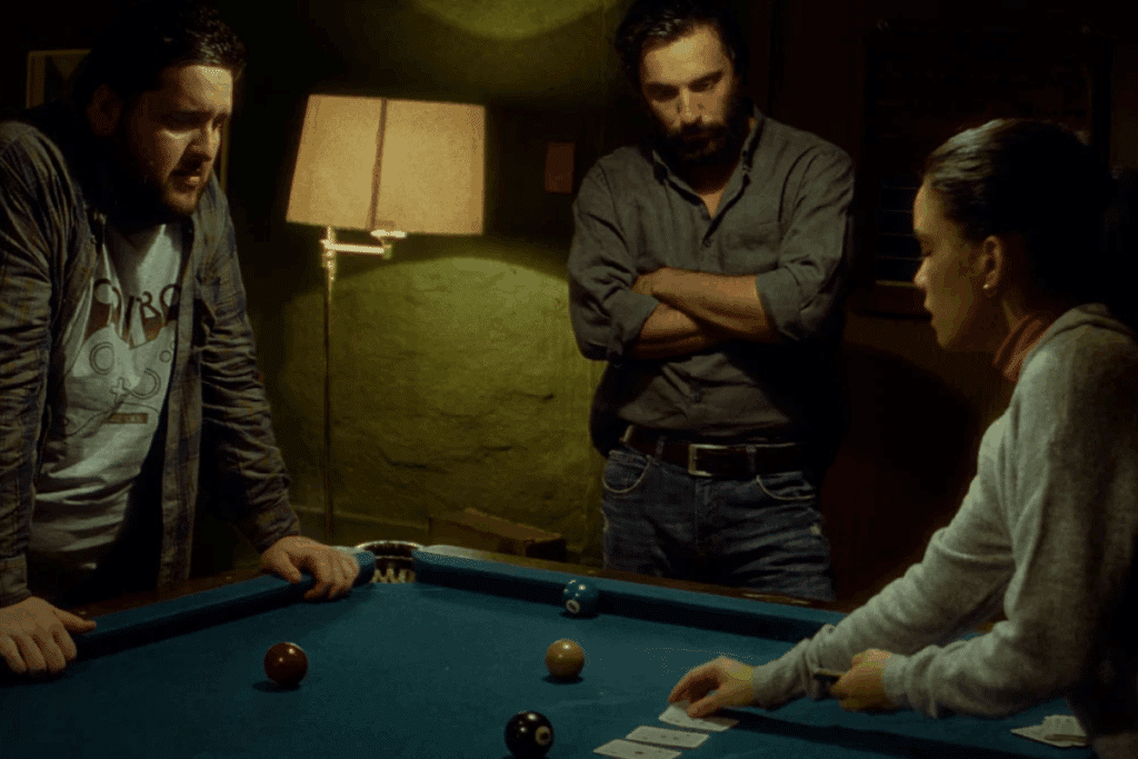 Três pessoas estão reunidas ao redor de uma mesa de sinuca azul sob iluminação penumbrosa. À esquerda, um homem inclina-se sobre a mesa; no centro, um homem de barba e braços cruzados observa atentamente; à direita, uma mulher distribui cartas de baralho sobre a superfície da mesa.