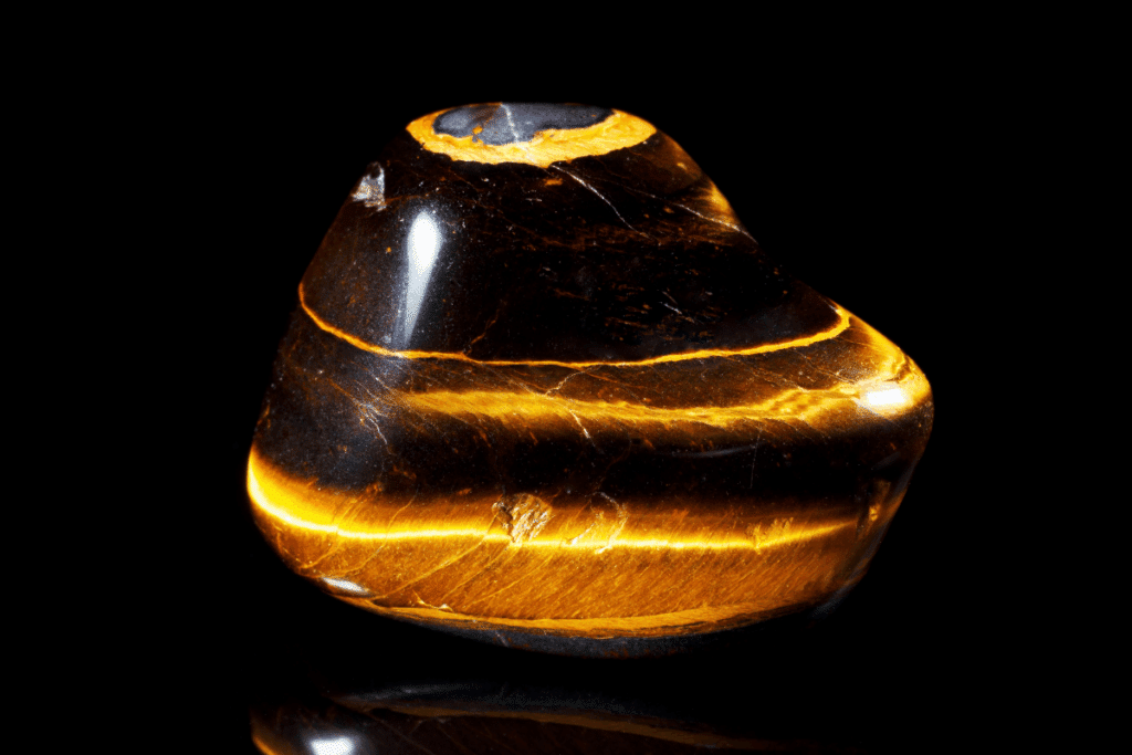 Pedra polida de olho-de-tigre com formato arredondado, destacando faixas horizontais brilhantes nos tons castanho e dourado.