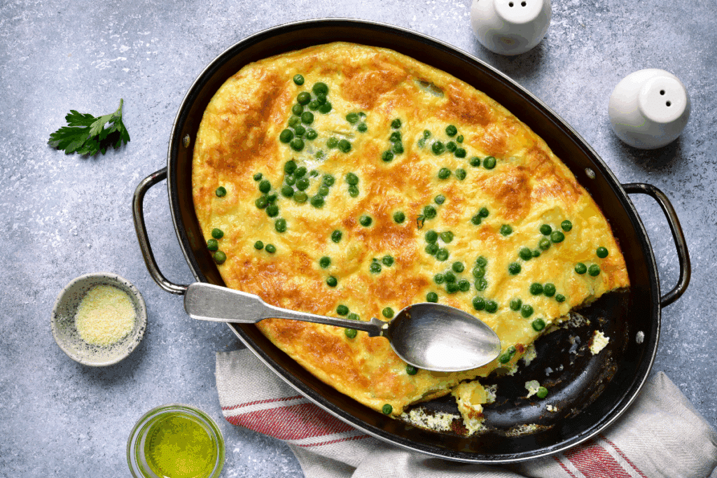 A imagem mostra um assado de ovos com ervilhas, preparado em uma assadeira oval escura. O prato apresenta uma superfície dourada e gratinada, com ervilhas verdes espalhadas por cima. Uma parte do assado já foi retirada, revelando o interior macio e cremoso.